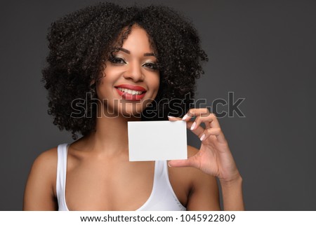 Young pretty African American millennial woman with natural curly hair wearing fashionable white tank top smiling holding a blank poster on a dark gray isolated background.
