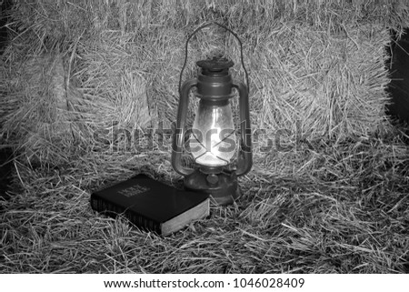 Black and white picture of a bible laying beside a oil lantern on a bale of hay.
