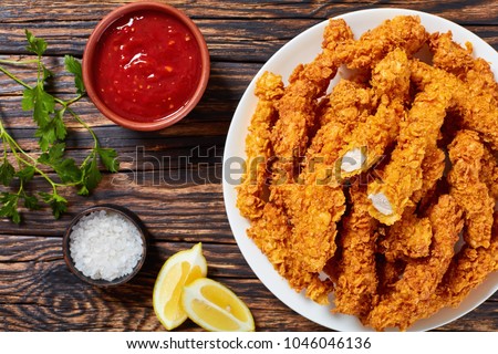corn flakes breaded deep fried chicken crispy breast strips on white plate, on old rustic  wooden table with tomato sauce and lemon slices, easy recipe for outdoor picnic or party, close-up Royalty-Free Stock Photo #1046046136