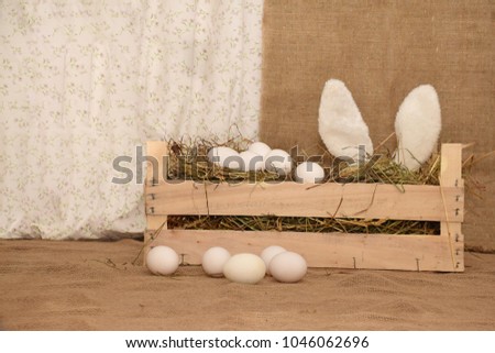 Easter picture. Hare ears peep out of a wooden box with hay. Next group of chicken white eggs.
