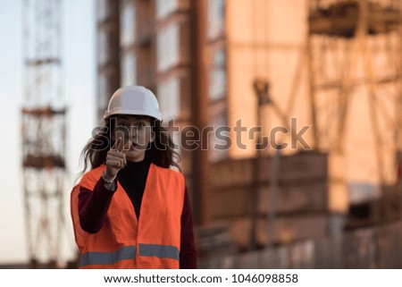 Beautiful young brunette in a helmet against the backdrop of the building. A girl wearing a protective orange vest and white helmet shows her finger straight. 
