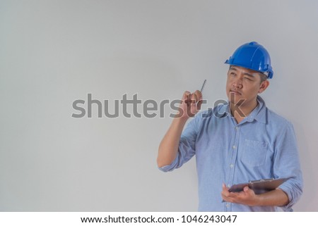 Thoughtful young man architect engineer or interior designer wear a blue safety helmet, he holding the pen and clipboard in his hand, with background factory wall.