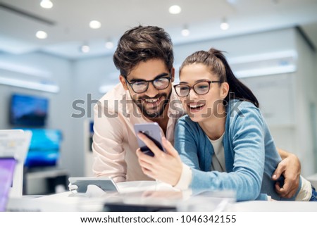 Young satisfied stylish charming couple testing the new model of a mobile from the desk in the tech store. Royalty-Free Stock Photo #1046342155