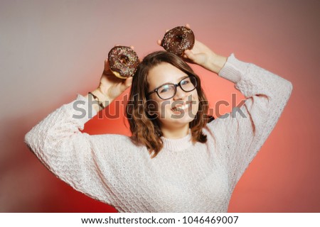 Happy young woman with eyeglasses having fun while holding donuts on head like ears and looking at the camera over pink background