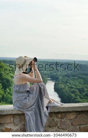 The girl in a sundress and a hat with a camera. Hill, green forest and river below