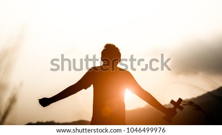 Young man holding christian cross and look up to the sky with light sunset background