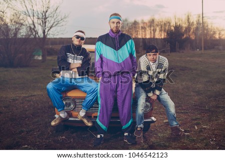 Three friends in the style of the nineties are about orange old car, Royalty-Free Stock Photo #1046542123