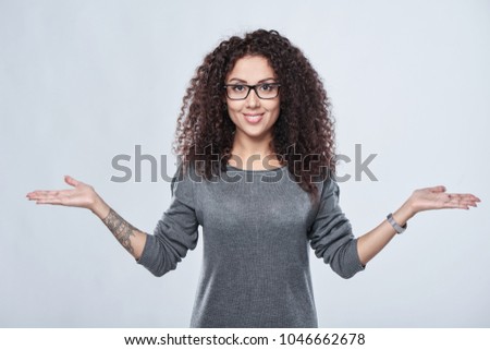 Closeup of smiling curly woman in eyeglasses with two opened hand palms with blank copy spaces contemplating choosing weighing, over grey background