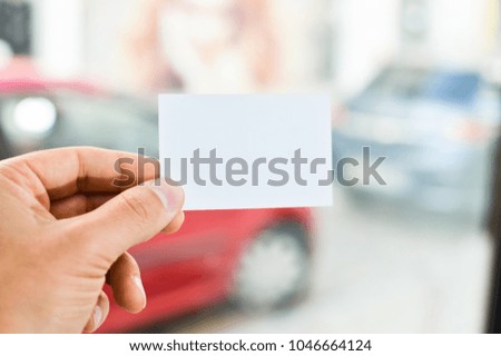 Close up on hand of a businessman holding white business card over blurry car office background. Professional person showing introduction