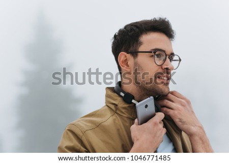 Portrait of handsome man looking down, wearing eyeglasses, coat and black headphones to listening the music from smartphone in the misty park outdoor. People, technology and lifestyle concept