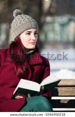 vertical photo young girl in hat, burgundy jacket and green pants, sits pondering with a book on a wooden bench in a winter park on a sunny day