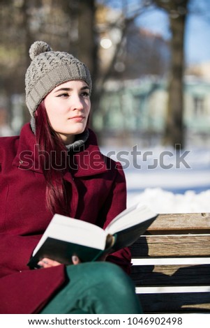 vertical photo of a young girl in hat, burgundy jacket and green pants, sitting with a book open on a wooden bench and staring into the distance in a winter park on a sunny day