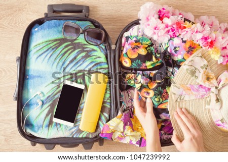 Opened travel bag with woman's flowery swimsuit, towel with palm tree picture, sun lotion and smartphone for relax in hot, exotic place. Preparation for traveling. Suitcase on wooden floor.