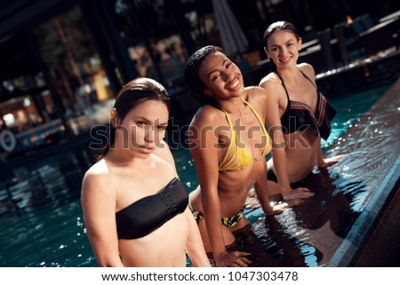 Three beautiful girls in swimsuits posing on the camera at swimming pool. Multiethnic company of friends in swimming pool.