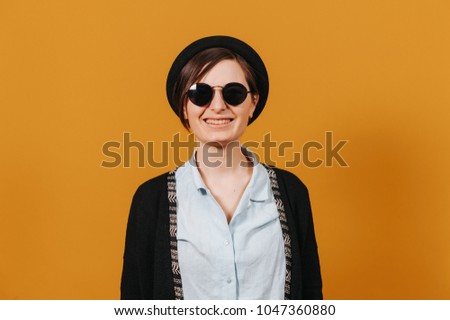 Young stylish brunette in black round sunglasses and hat smiling at camera on orange backdrop. 