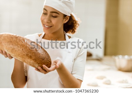 smiling female baker holding fresh loaf of bread on baking manufacture