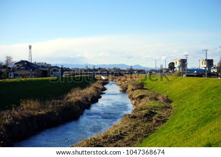 A landscape of river with green grass, small house, and background mountain in suburban under the blue sky