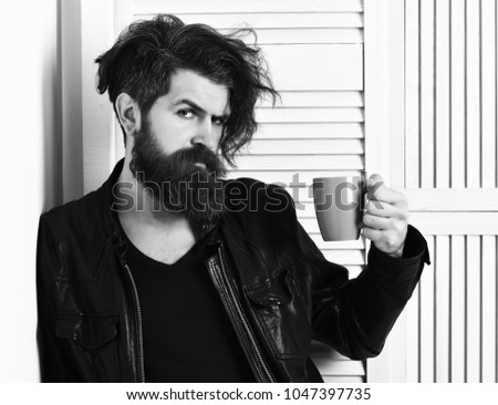 Bearded man, long beard. Brutal caucasian hipster with moustache and ruffled hair holding cup or mug in rock black style on white wooden vintage studio background
