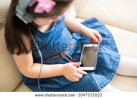 Top view of little girl sitting sofa and watching cartoon movie on mobile phone with headphones on
