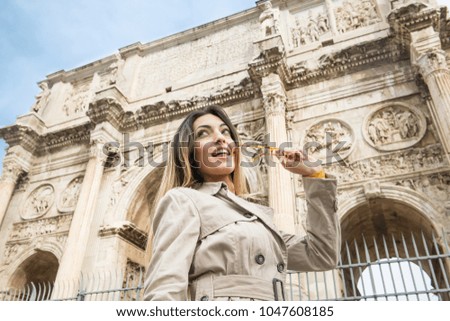 Low angle view of young pretty tourist woman smiling looking out eyeglasses in mouth, in background arch of Constantine monument in Rome Italy