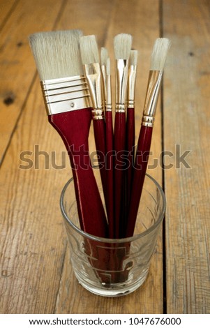Bristle paint brushes with red rods in glass cup on wood background