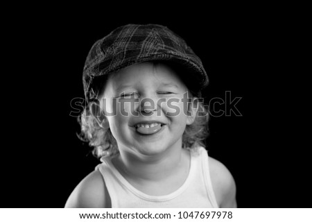 A black and white portrait of a little boy wearing a white tank top, sticking out his tongue. The child's eyes are closed and his tongue is out. There is room for tex on the solid black background.