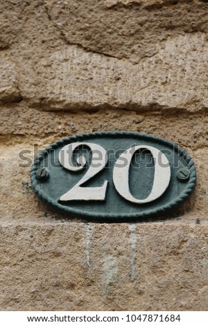 Close up outdoor view of the number twenty written in white on a blue oval metallic plate. Element fixed on a stone bricks wall of a french house to indicate the address. Decorative sign in the street