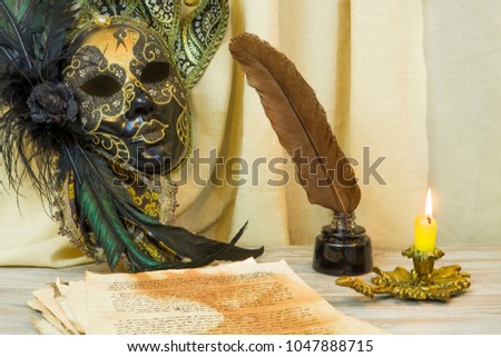 Literature concept, candle in a candlestick, near a Venetian mask and an inkwell with a pen on a background of bright material