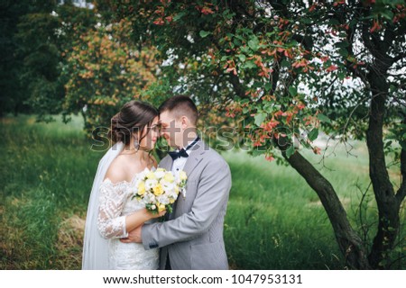 Stylish newlyweds stand forehead to the forehead and gently embrace, against the backdrop of beautiful trees and red flowers.
