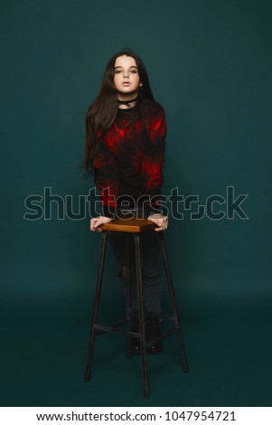 Young brunette teen girl stands near the chair over dark-green background, isolated in studio
