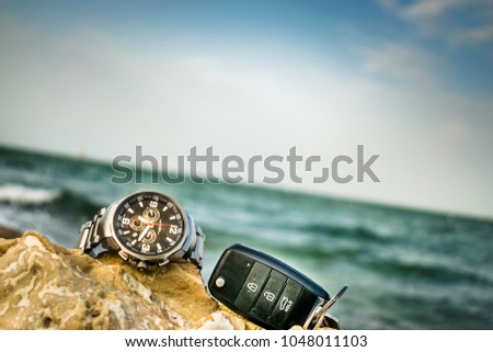 a big dial silver watch kept on rocks with a modern remote car key and greenish see in the background 