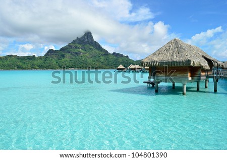 Luxury overwater bungalow with a thatched roof in a honeymoon vacation resort in the clear blue lagoon with a view on Mt. Otemanu on the tropical island of Bora Bora, near Tahiti, in French Polynesia. Royalty-Free Stock Photo #104801390