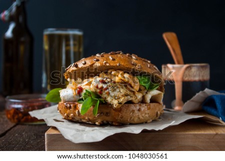 
Burger with fish with spicy sauce, lamb's lettuce, sun-dried tomatoes and onions. Served on a wooden board. Beer in the background.
