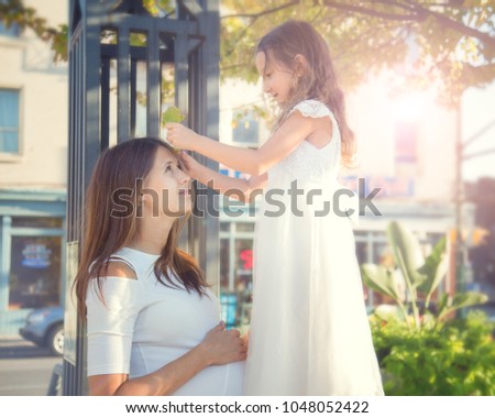 Little cute girl putting leaf in her pregnant mother's long hair. Sunny day. Mother's day
