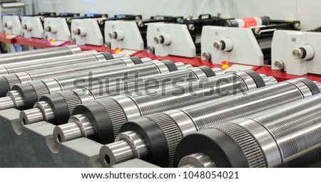 Lined up magnetic cylinders for die cut on rotary printing press. Magnetic cylinder for flexo rotary die cutting. Magnetic roll and in-line press machine in background. Cylinder for cutting dies. Royalty-Free Stock Photo #1048054021