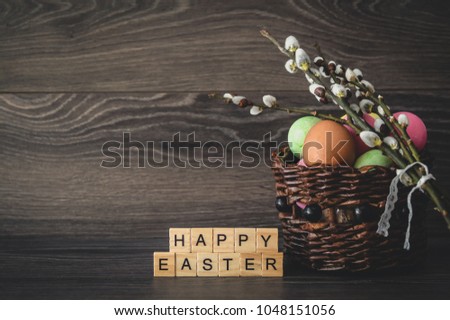 The inscription  happy Easter, made of light wooden cubes, painted eggs in a wicker basket and willow branches with seals on a dark wooden background