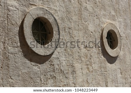 Close up outdoor urban view of a circular window in a grey concrete wall. Isolated element with glass and railings. Geometric shape with its shadow. Rough texture. Abstract architectural image.