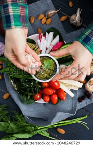 Woman hand adds sesame seeds to healthy veggie buddha bowl with vegetables - cucumbers, tomatoes, radish, mushrooms, seaweed, garlic,  avocado chickpea dip. Above top view. Raw, vegan, vegetarian food