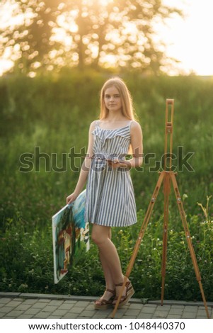 Beautiful girl draws a picture in the park using a palette with paints and a spatula. Easel and canvas with a picture.