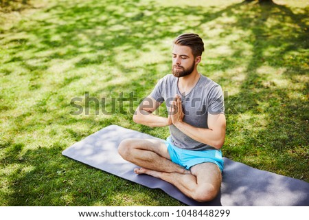 Young man meditating outdoors in the park, sitting with eyes closed and his hands together Royalty-Free Stock Photo #1048448299
