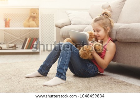Little girl playing online games on tablet and and hugging toy. Female child sitting on the floor near sofa with her teddy bear. Kid leisure at home. Social networking and online education concept