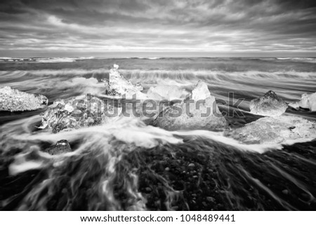 Ice blocks lie on a black beach, the movement of the waves can be seen (long exposure), above a contrasting cloudy sky - Picture in black and white - Location: Iceland, Jökulsárlón (Jökulsarlon)