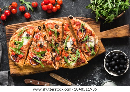 Flatbread pizza garnished with fresh arugula on wooden pizza board, top view. Dark stone background Royalty-Free Stock Photo #1048511935
