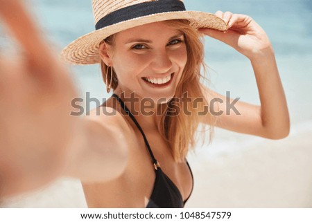 Close up shot of good looking female tourist enjoys free time outdoor near ocean on beach, looks at camera during leisure on sunny summer day, poses for selfie. Happy smiling tourist in tropics