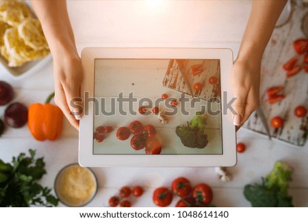 View of female hands holding white digital tablet with colorful photo on the screen of healthy salad recipe with vegetables. Selective focus on touch pad on which female food blogger taking picture.