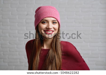 Beautiful positive model in red hat and sweater smiling to camera while posing in front of blurred white brick wall in professional photo studio. Horizontal shot.