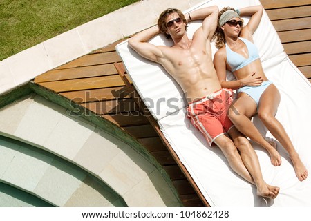 Over head view of a young, attractive couple sunbathing by a swimming pool while on holiday.