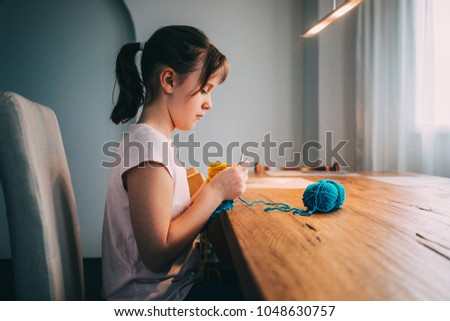 Young girl knitting a circle scarf with yellow and blue coloured yarn. Sitting at the wooden table, side view.