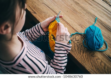 Young girl knitting a circle scarf with yellow and blue coloured yarn. Sitting at the wooden table, close up over the shoulder.