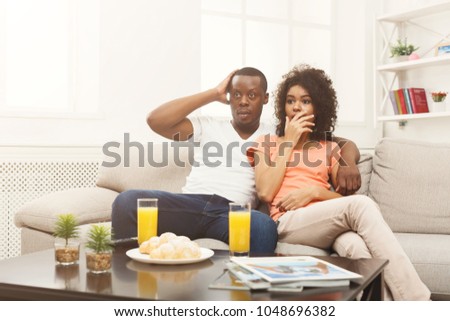Surprised and frightened african-american couple relaxing and watching TV at home, having rest after hard week, copy space